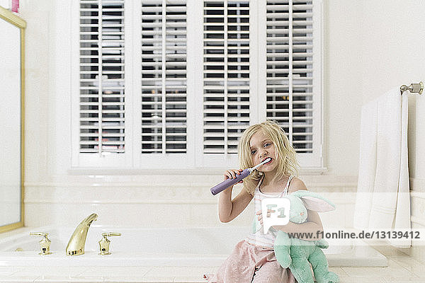 Cute girl brushing teeth while holding stuffed toy at bathroom