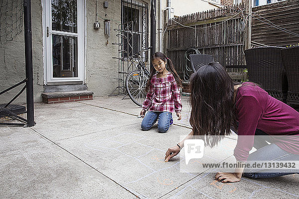 Girl looking at sister drawing hopscotch outside house