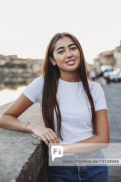 Porträt einer lächelnden jungen Frau  die an einer Stützmauer in der Stadt steht