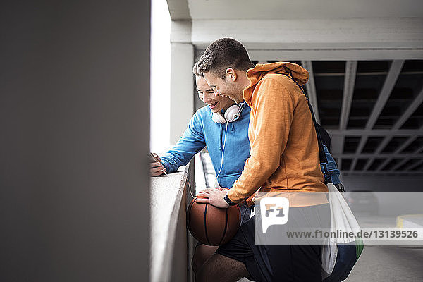 Fröhliche männliche Freunde mit Basketball mit Smartphone auf dem Parkplatz