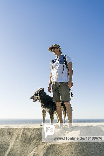Mann mit Hund steht am Strand vor klarem blauen Himmel