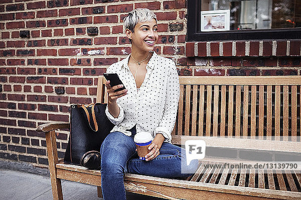 Woman looking away while holding mobile phone and coffee cup on bench