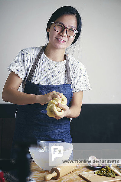 Porträt einer selbstbewussten Frau  die Teig knetet  während sie in der Küche an der Wand steht