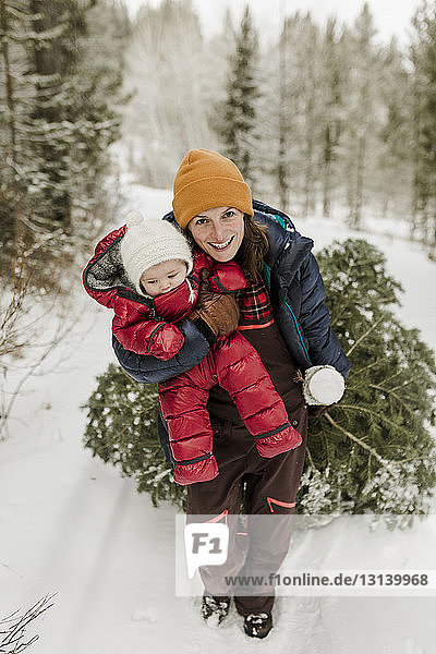 Porträt einer lächelnden Mutter  die eine Tochter trägt  während sie im Winter eine Kiefer im Wald zieht