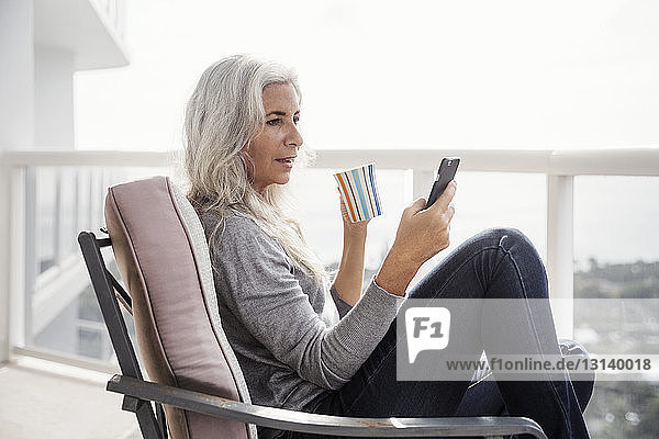 Seitenansicht einer reifen Frau  die ein Smartphone benutzt  während sie auf dem Balkon Kaffee trinkt