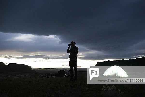 Silhouette hiker photographing while standing on field by tent against stormy clouds during dusk