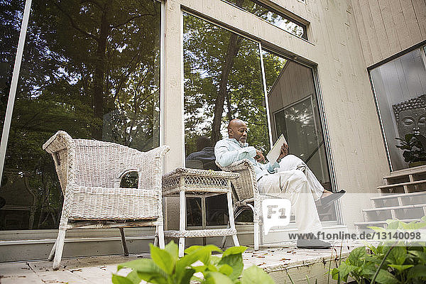Älterer Mann benutzt Tablette  während er vor dem Haus auf einem Korbstuhl sitzt