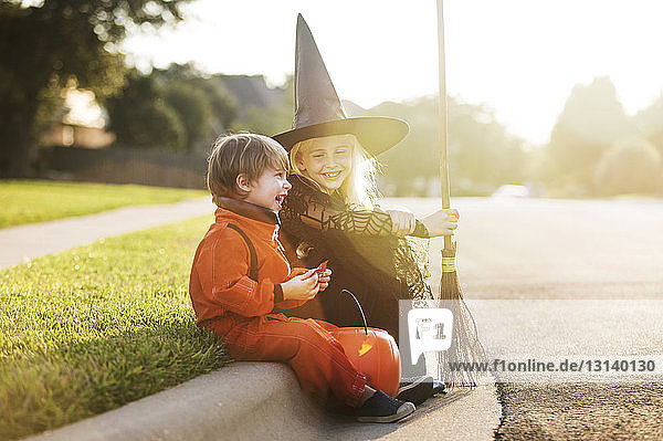 Fröhliche  für die Halloween-Party gekleidete Geschwister sitzen an der Straße