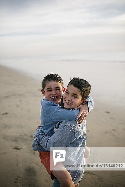 Portrait of boy carrying happy brother while standing at beach