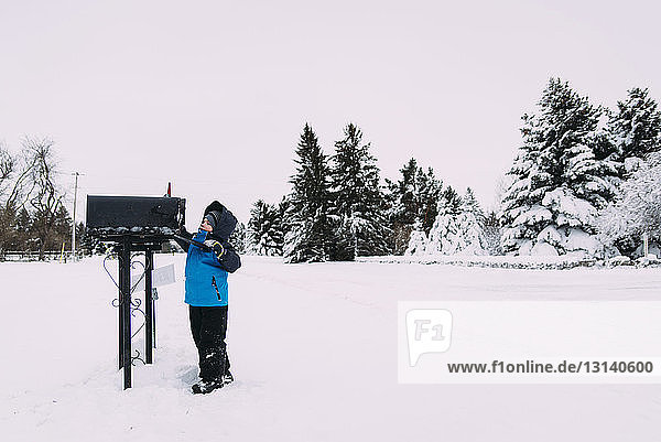 Junge in warmer Kleidung steht auf schneebedecktem Feld mit Briefkasten vor klarem Himmel