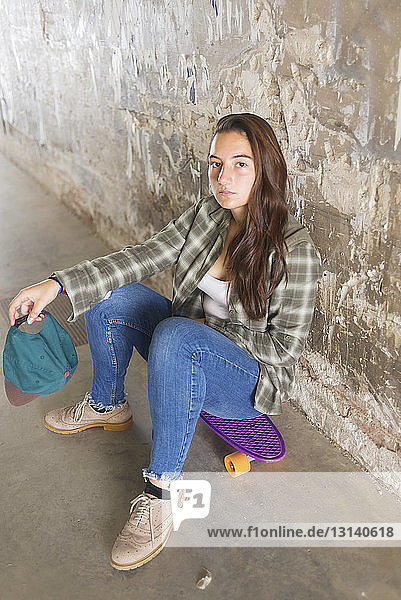 Portrait of confident student sitting on skateboard against weathered wall