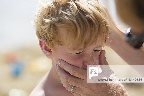 Geschnittene Hände eines Vaters  der im Sommer am Strand das Gesicht seines Sohnes hält