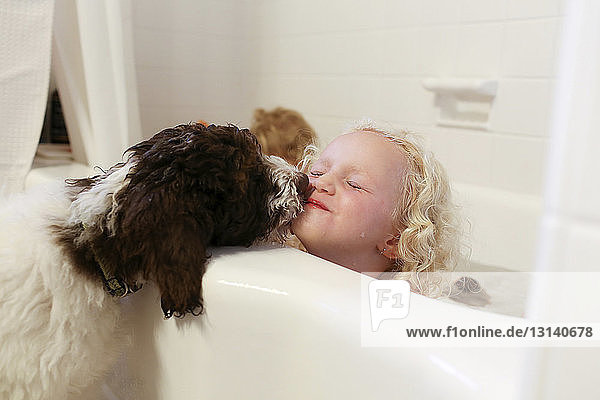 Dog kissing girl sitting in bathtub in bathroom