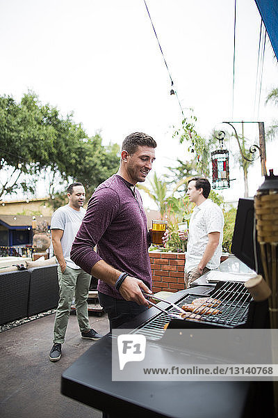 Mann bereitet Essen auf dem Grill zu  während er mit Freunden im Hintergrund steht