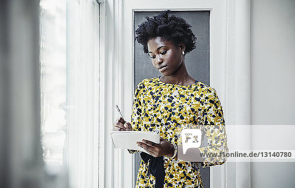 Geschäftsfrau schreibt in Buch  während sie im Kreativbüro am Fenster steht