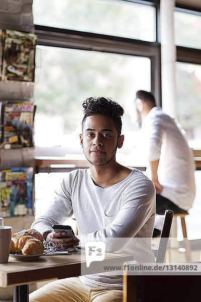 Portrait of young man with mobile phone having croissant while sitting at cafe