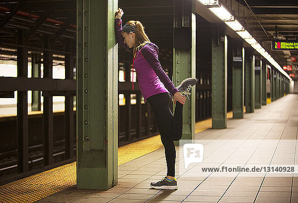 Seitenansicht einer jungen Frau  die sich am Bahnhof an einer Säule lehnend streckt