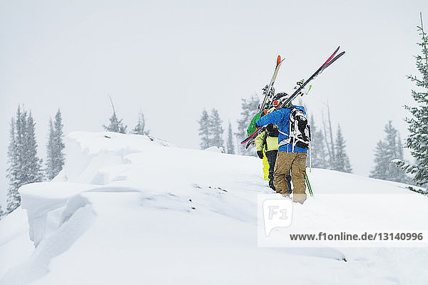 Rear view of friends carrying skis while walking on snow covered mountain