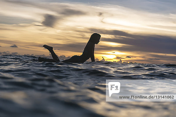 Seitenansicht der Silhouette einer jungen Frau  die bei Sonnenuntergang im Meer gegen einen bewölkten Himmel schwimmt
