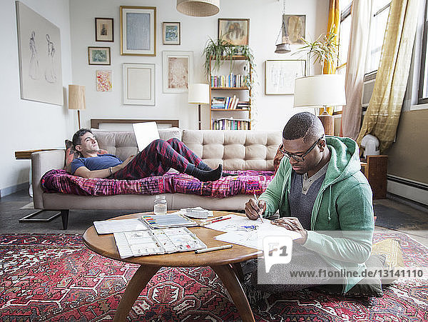 Homosexueller Mann zeichnet  während die Partnerin den Laptop zu Hause benutzt