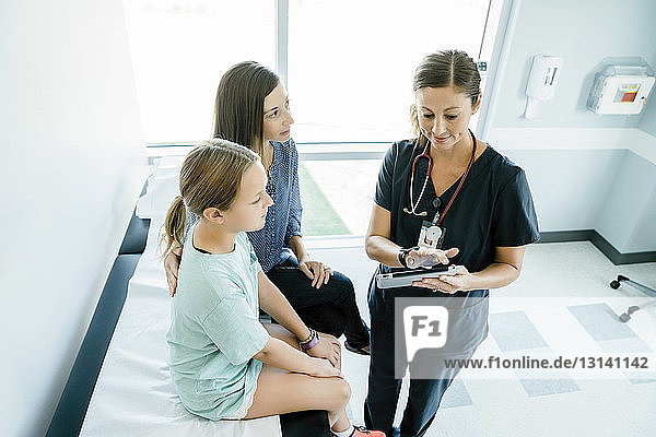 Hochwinkelansicht eines Kinderarztes  der Mutter und Tochter  die auf dem Untersuchungstisch sitzen  einen Tablet-Computer zeigt