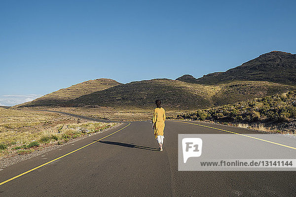 Rückansicht einer Frau  die bei strahlend blauem Himmel an einem sonnigen Tag auf einer Straße inmitten von Bergen geht