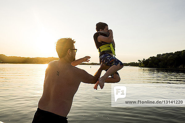 Rückansicht eines hemdlosen Vaters  der seinen Sohn bei Sonnenuntergang gegen den klaren Himmel in einen See wirft