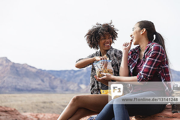 Female friends eating while sitting on rock formation against clear sky during sunny day