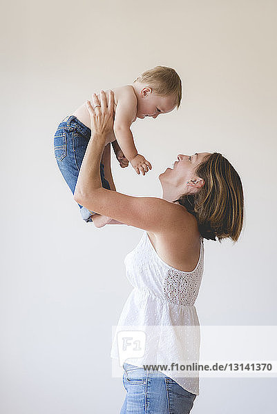 Side view of playful mother lifting shirtless son against white background