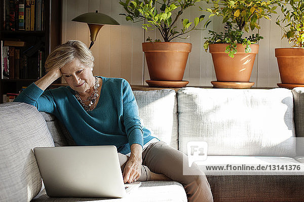 Frau mit der Hand im Haar  die einen Laptop benutzt  während sie zu Hause auf dem Sofa sitzt