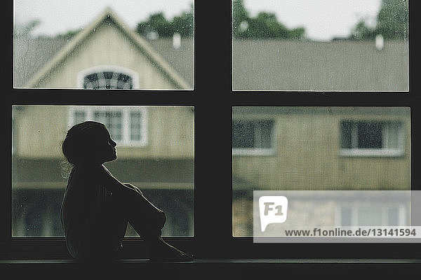 Thoughtful girl sitting on window sill against house