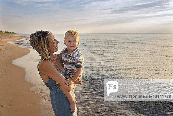 Porträt eines süßen Sohnes  der von der Mutter bei Sonnenuntergang am Strand getragen wird
