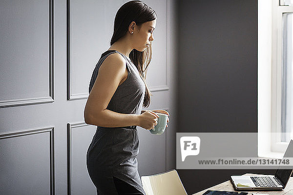 Businesswoman with mug looking at laptop computer while standing in office