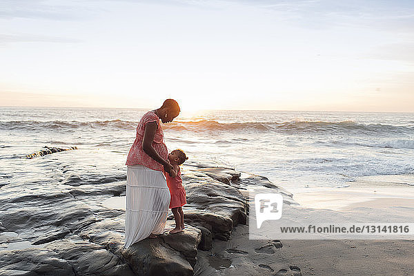 Tochter küsst schwangeren Mutterbauch  während sie am Strand gegen den Himmel steht