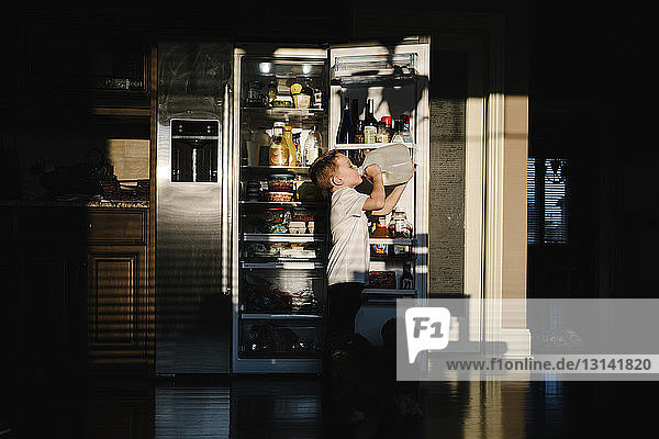 Seitenansicht eines Jungen  der Milch aus einer Flasche trinkt  während er zu Hause in der Dunkelkammer gegen einen offenen Kühlschrank steht