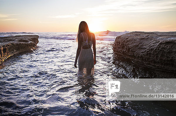 Rückansicht eines Teenagers  der bei Sonnenuntergang an einer Felsformation im Meer steht