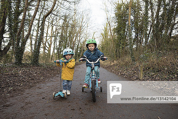 Portrait of siblings riding push scooter and bicycle on road against sky