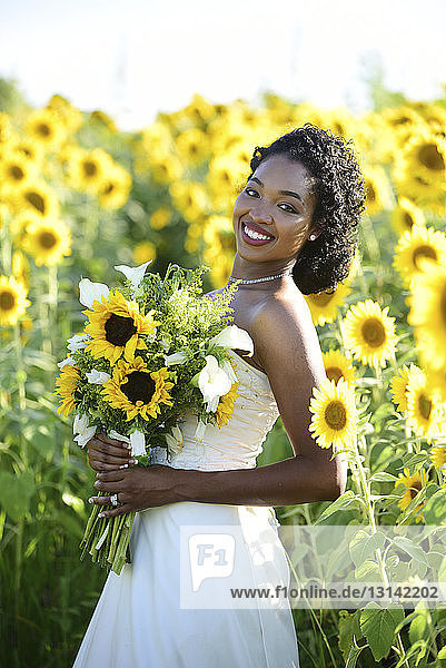 Porträt einer lächelnden Braut  die einen Blumenstrauss hält  während sie inmitten von Sonnenblumen auf dem Bauernhof steht