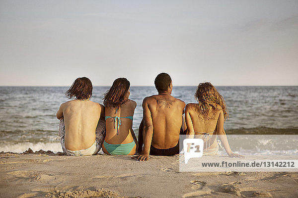 Rückansicht von am Strand sitzenden Freunden gegen den Himmel