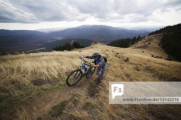 Male backpacker climbing with bicycle on mountain against cloudy sky