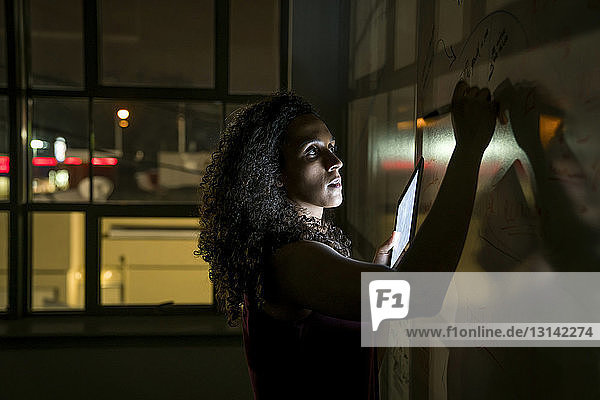 Geschäftsfrau schreibt in dunklem Büro auf Whiteboard über Tablet-Computer
