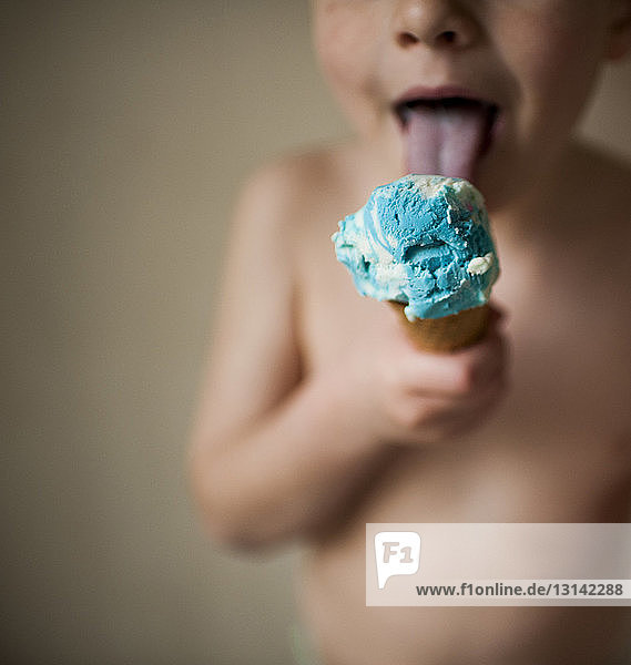 Midsection of shirtless boy licking ice cream while standing against wall at home
