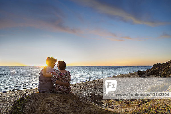 Rückansicht eines Paares  das bei Sonnenuntergang auf einem Felsen am Strand gegen den Himmel sitzt