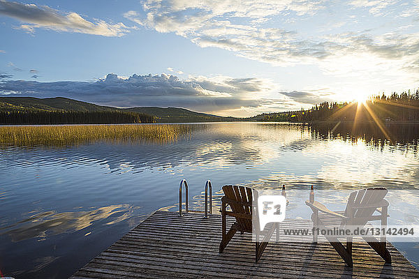 Leere Stühle auf Holzsteg über dem See vor bewölktem Himmel bei Sonnenuntergang