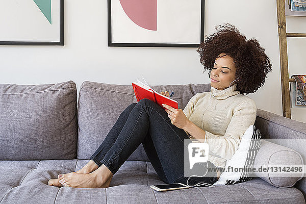 Frau schreibt in Buch  während sie über ein Smartphone auf dem Sofa Musik hört