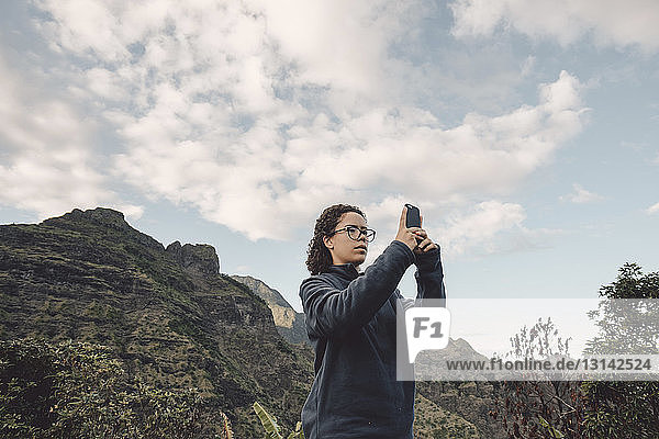 Low angle view of woman photographing with mobile phone while standing against mountain
