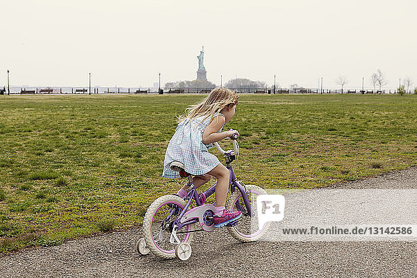 Side view of girl riding bicycle on road by field with Statue of Liberty in background