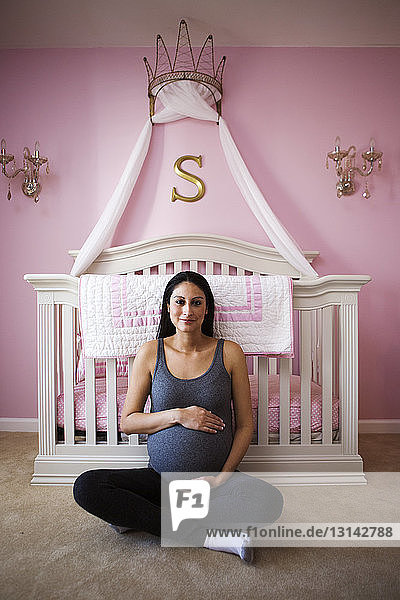 Porträt einer lächelnden schwangeren Frau  die auf dem Boden vor der häuslichen Krippe sitzt
