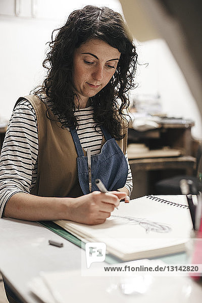 Female artisan drawing on book while sitting in workshop