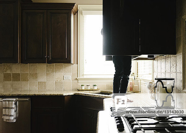 Unterer Teil eines Jungen  der zu Hause auf der Küchentheke steht und etwas im Schrank sucht
